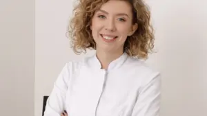 Šviesiaplaukė garbanota gydytoja baltais chalatais laiko apsauginius akinius ir šypsosi žiūrėdama į kamerą.