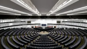 Tuščia Europos Parlamento posėdžių salė su puslankiu išdėstytomis darbo vietomis.