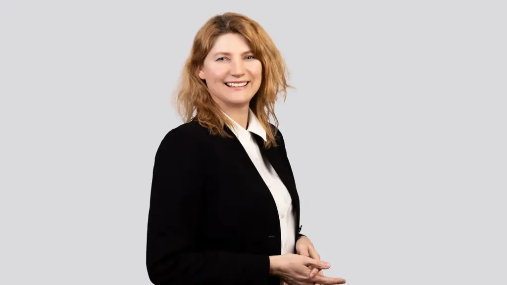 Besišypsanti šviesiaplaukė moteris vilkinti baltus marškinius ir juodą švarką šviesiame fone.